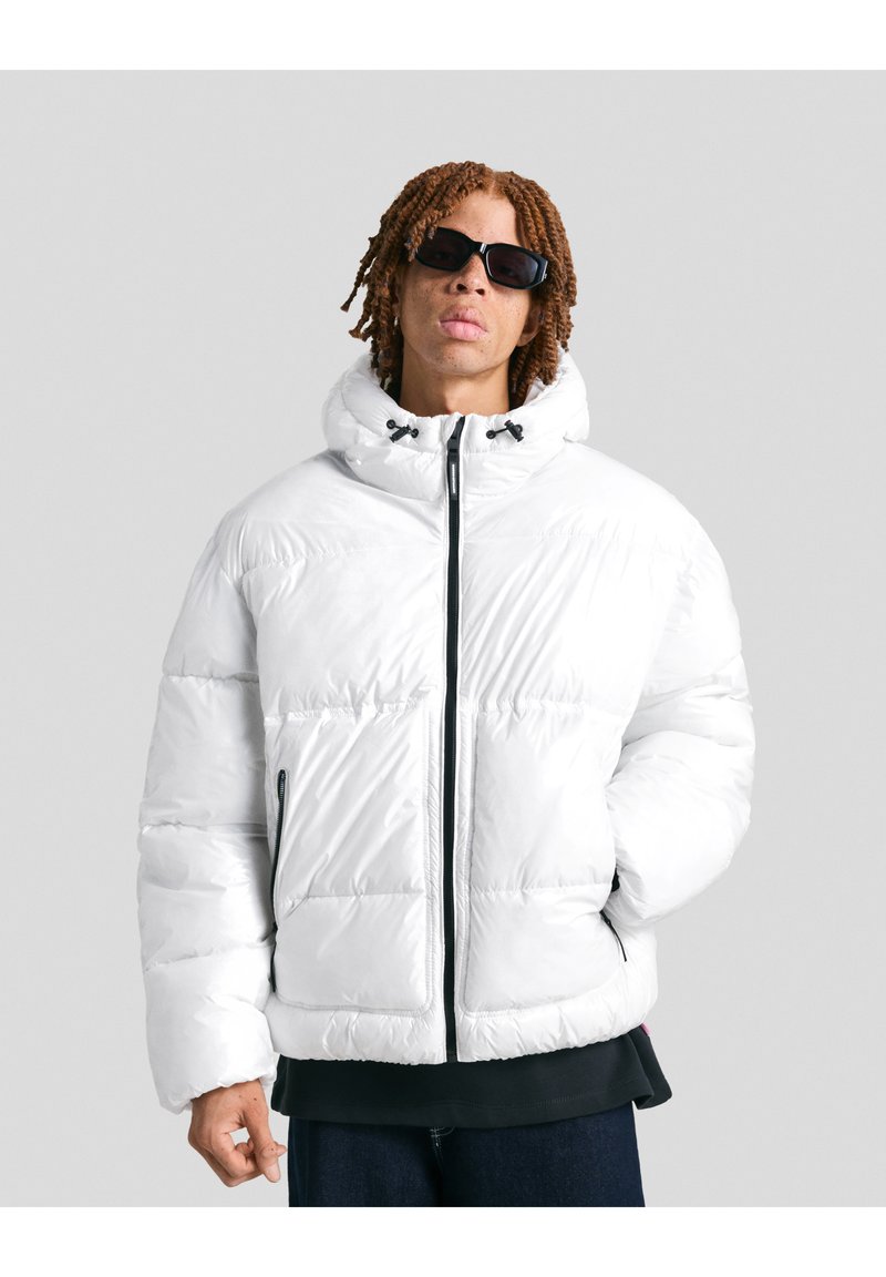 Persoană cu părul scurt și creț, purtând ochelari de soare negri și o jachetă albă din puf cu glugă, stând în fața unui fundal gri uniform.