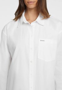 Camicia bianca con colletto classico, taschino sul petto e tessuto liscio e leggero. Design semplice, senza motivi visibili.