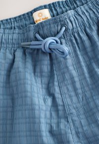 Vaaleansiniset uimalaukut, jotka on valmistettu teksturoidusta kankaasta. Niissä on joustava vyötärönauha, jossa on kiristysnauha, ja niissä on etiketti, jossa lukee "Next Swim".