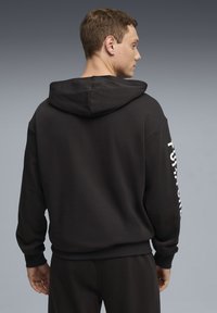 Sweat-shirt à capuche noir avec une coupe décontractée. Comprend des poignets et une ceinture côtelés, ainsi qu'un texte blanc audacieux sur la manche gauche. Texture douce.