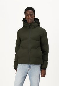 Mørkegrøn pufferjakke med hætte, der har en frontlynlås og elastiske manchetter. Bæret over lyseblå jeans, glat tekstur og let glinsende finish.