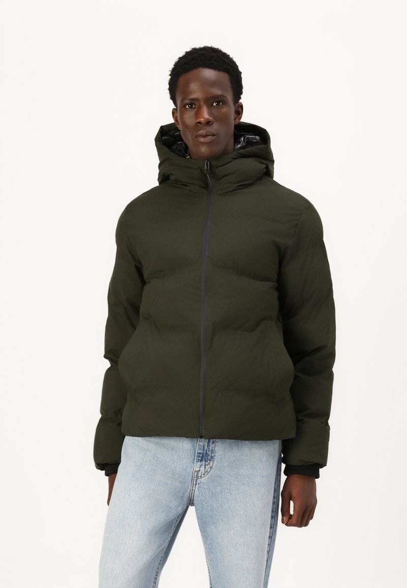 Mørkegrøn pufferjakke med hætte, der har en frontlynlås og elastiske manchetter. Bæret over lyseblå jeans, glat tekstur og let glinsende finish.