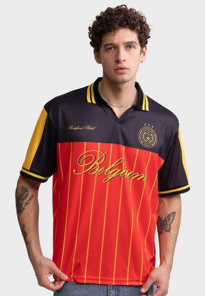 Young man with curly hair wearing a black and red Belgium sports jersey with yellow stripes, showing tattoos on both forearms.