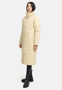 Beige lange Daunenjacke mit hohem Kragen, Seitentaschen und Reißverschluss. Getragen über dunklen Leggings und schwarzen Ankle Boots. Glatte Textur, minimalistisches Design.