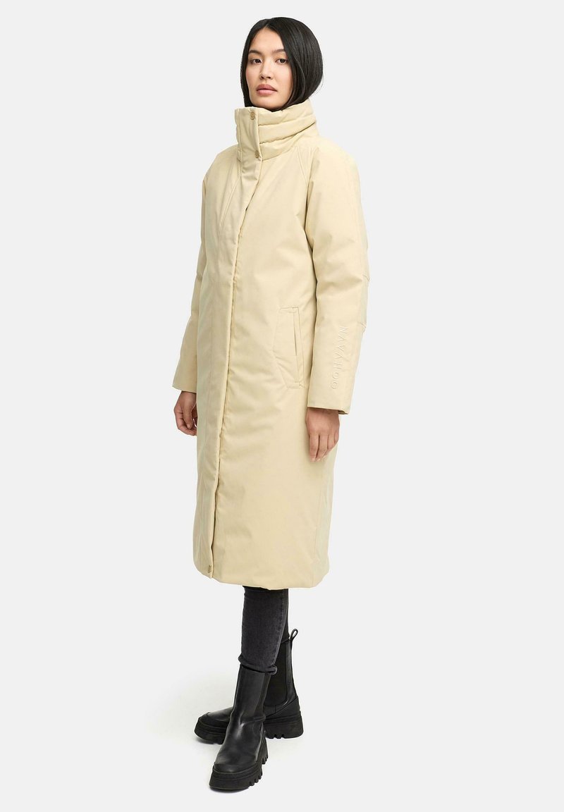 Beige lange Daunenjacke mit hohem Kragen, Seitentaschen und Reißverschluss. Getragen über dunklen Leggings und schwarzen Ankle Boots. Glatte Textur, minimalistisches Design.