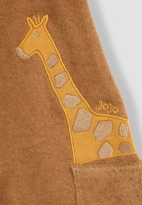 Tela marrón con un bordado de jirafa en amarillo y beige. La jirafa tiene manchas y un pequeño ojo azul claro. Logo "Jojo" cerca del diseño.