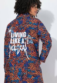 Kleurrijke, gestreepte trenchcoat met een luipaardprint in oranje, blauw en zwart. Bevat witte tekst "LIVING LIKE A ROCKSTAR" en steraccenten.