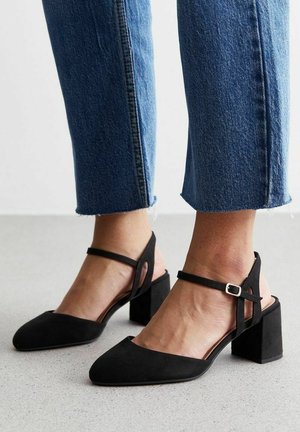 Fødder iført sorte ruskindssko med blokhæl og ankelremme, kombineret med cropped blå denim jeans mod en grå gulvbaggrund.