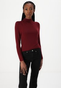 Burgundy långärmad topp med hög hals och subtila puffärmar, kombinerad med svarta jeans. Slät textur och åtsittande design.