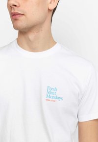 Witte katoenen t-shirt met korte mouwen, met "Fresh Meat Mondays" in het blauw en "REVOLUTION™" in het oranje op de linkerkant van de borst.
