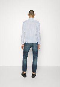 Man som står vänd bort, iförd en vit mönstrad skjorta med knappar, blå jeans och svarta skor framför en helt vit bakgrund.