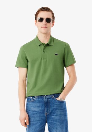 Groen poloshirt met kraag, korte mouwen en een klein zwart logo op de borst, gecombineerd met blauw denim jeans en een zonnebril.
