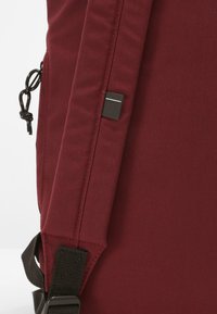 Mochila de tela burdeos con superficie texturizada, correa negra ajustable y un pequeño acento metálico en el lateral.