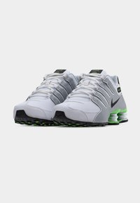 Παπούτσια Nike Shox σε λευκό και γκρι με μαύρες λεπτομέρειες, με υφασμένο επάνω μέρος, χαρακτηριστική απορρόφηση κραδασμών Shox και πράσινες λεπτομέρειες στην εξωτερική σόλα.