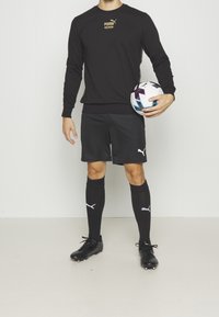 Felpa sportiva nera con logo dorato, pantaloni corti neri, calze nere con logo bianco e scarpe da calcio nere, mentre tiene un pallone da calcio multicolore.