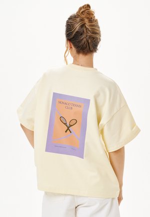 Kobieta z włosami upiętymi w kok, nosząca kremowy, luźny t-shirt z fioletowo-pomarańczowym motywem tenisowym na plecach.
