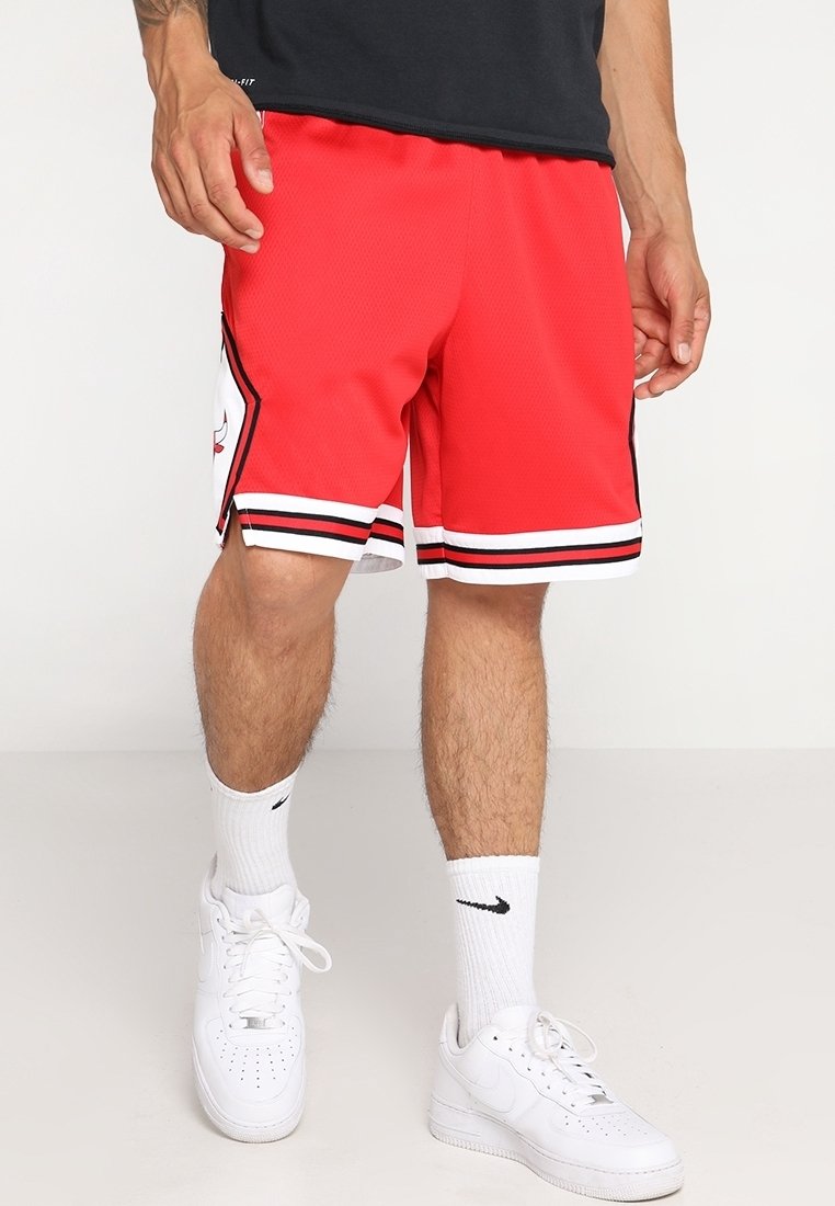 Homme portant un short de basket rouge avec des rayures noires et blanches, des chaussettes blanches Nike et des baskets blanches Nike, debout sur un fond uni.