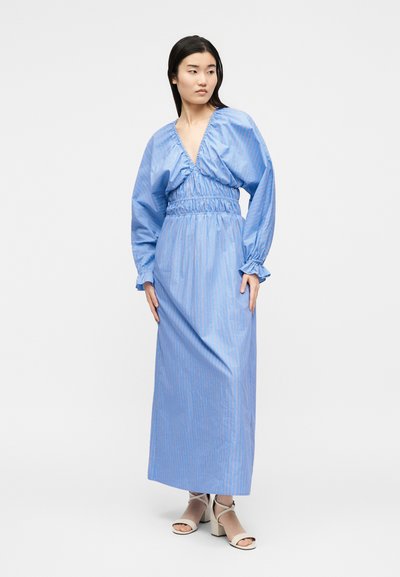 Γυναίκα που φοράει μακρύ ανοιχτό μπλε φόρεμα με φουσκωτά μανίκια και σούρες στη μέση, στέκεται με τα χέρια χαλαρά, κοιτάζοντας στο πλάι, ορατά λευκά σανδάλια.