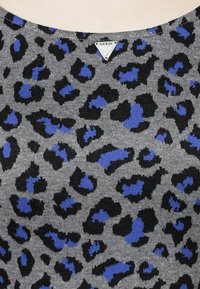 Top en tricot gris avec un motif léopard noir et bleu, orné d'une étiquette en métal triangulaire 'Guess' au niveau du col.