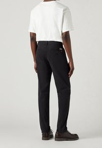 Personne portant une chemise blanche à manches courtes, un pantalon noir avec des poches arrière boutonnées et des chaussures marron foncé à lacets, debout devant un fond uni.