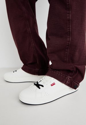 Hvide lave sneakers med sorte snørebånd og rød Levi's-seddel, båret med mørkerøde bukser, stående på en hvid overflade.
