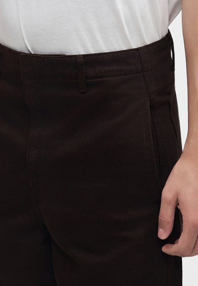 Gros plan d'une personne portant un pantalon marron foncé et une chemise blanche, montrant la ceinture et la main droite reposant près de la hanche.