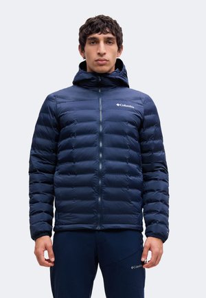 Jeune homme portant une doudoune bleu marine Columbia avec capuche et un pantalon bleu marine assorti, debout face à l'avant avec une expression neutre.