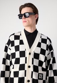 Κασκόλ σε ασπρόμαυρη καρώ διάταξη με V-λαιμό, κουμπί κλεισίματος και μπροστινή τσέπη που φέρει το λογότυπο "HUGO", φορεμένο πάνω από μαύρο polo.