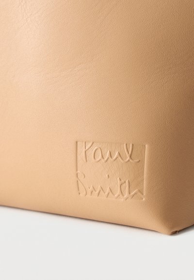 Surface en cuir beige avec un logo rectangulaire embossé portant l'inscription "Paul Smith" en écriture cursive.