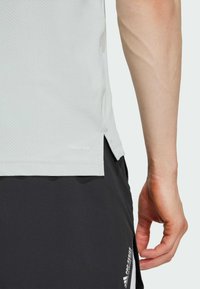 Camiseta deportiva de color gris claro con un acabado texturizado y aberturas laterales, con un logo cerca del dobladillo. Combinada con pantalones cortos negros.