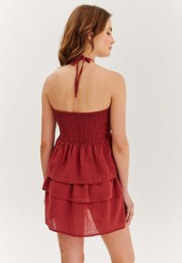 Žena s vlnitými vlasmi, oblečená v bezrukávových, tierovaných červených šatách s naťahovacou zadnou časťou a zaväzovaním za krkom, tváriaca sa od obrazu na jednoduchom pozadí.