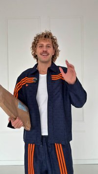 Fato de treino azul-marinho com riscas laranja; tecido texturado; modelo segurando um pacote de papel castanho; de pé contra um fundo branco liso.