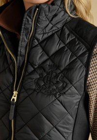 Zbliżenie osoby w czarnej, pikowanej kamizelce z złotym zamkiem i haftowanym logo na piersi, noszącej brązową bluzkę w wzór houndstooth.