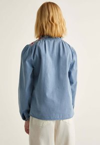 Cămașă din denim albastru cu mâneci lungi, detalii plisate la umeri și broderie florală de-a lungul decolteului. Textură netedă și croială lejeră.