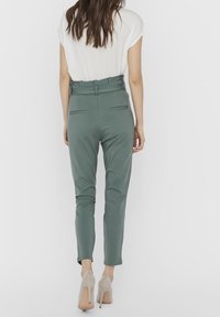 Groene aansluitende broek van glad materiaal, met een hoge tailleband, zijzakken en een taps toelopend ontwerp bij de enkels, gecombineerd met lichte hakken.