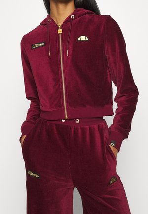 Vrouw in een bordeauxkleurige velours zip-up hoodie en bijpassende broek met ellesse-logo's, handen in de zakken, cropped bij de hals en midden boven de dijen.