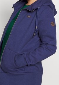 Marineblauwe zip-up hoodie gemaakt van zachte stof, voorzien van een capuchon met trekkoord, zijzakken en een logo patch op de mouw.