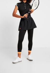 Kvinna i svart träningsoutfit som håller en tennisracket och boll, iklädd vita sneakers med orangea detaljer på leggings.