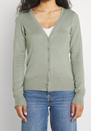 Cardigan verde chiaro con scollo a V, maniche lunghe e chiusura a bottoni. Tessuto morbido, polsini e vita a coste. Indossato sopra un top bianco.