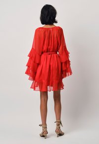 Rotes Chiffonkleid mit geschichteten, rüschigen Ärmeln, tailliertem Bund und ausgestellt Rock. Kombiniert mit nudefarbenen Riemchenabsätzen. Sichtbare Rückenverarbeitung.