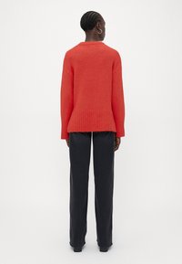 Maglione rosso lavorato a maglia con orlo e polsini a coste, abbinato a pantaloni neri a gamba larga. Il maglione ha una vestibilità comoda e una parte posteriore leggermente più lunga.