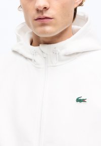 Νεαρό άτομο φορεί λευκό φούτερ με φερμουάρ και κουκούλα, με μικρό πράσινο λογότυπο κροκόδειλου στο στήθος.