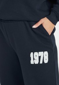 Hand i mörkblå sweatpantsficka med ett vitt chenille-märke med "1978" på låret, vilket visar detalj av avslappnad klädsel.