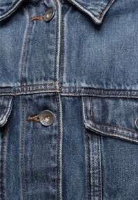 Veste en jean bleue avec des boutons métalliques et des coutures orange visibles le long des coutures et des poches.