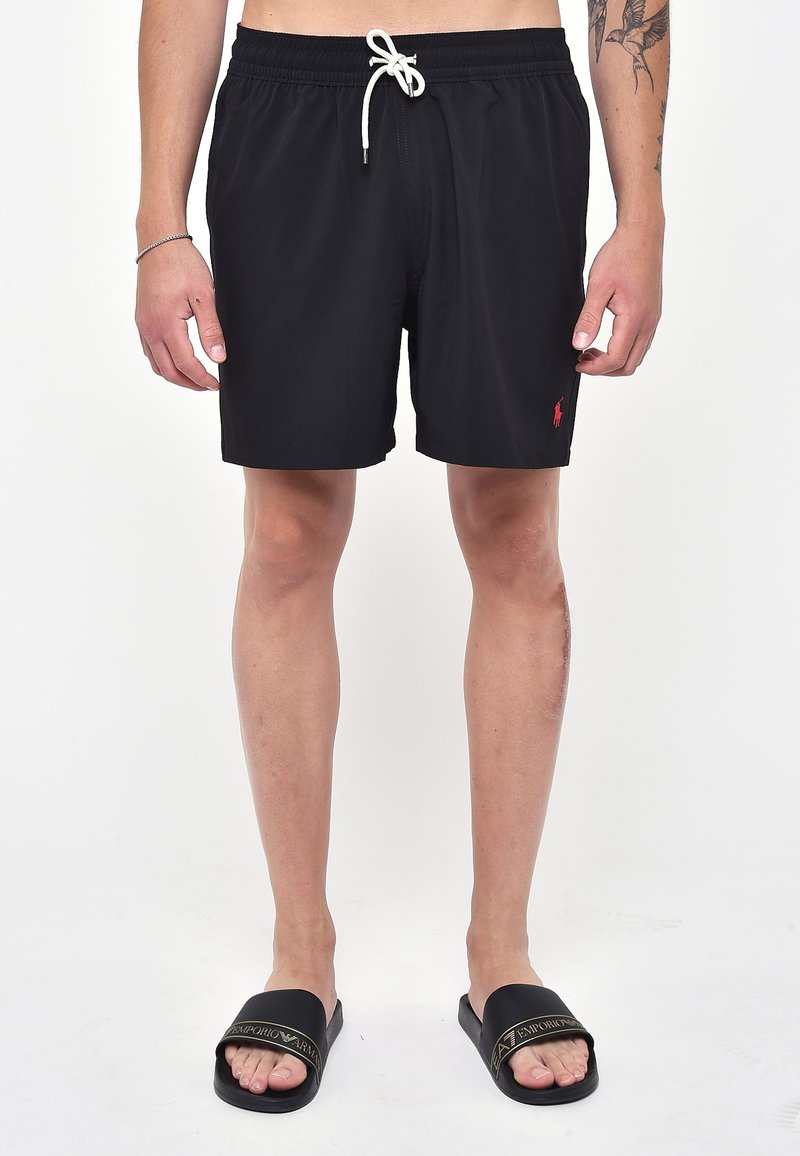 Pantaloni da bagno neri con cintura elastica, coulisse bianca e piccolo logo rosso. Indossati con ciabatte nere con scritta dorata.