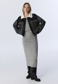 Vrouw in een grijze gebreide crop top en rok met een zwart en grijs jack met witte kraag en zwarte kniehoge laarzen, staand met handen op de heupen.