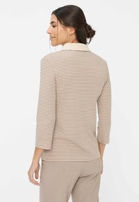 Beige und creme gestreiftes Langarmoberteil mit Kragen, das eine lockere Passform und weiche Textur aufweist. Getragen mit beigen Hosen.