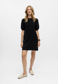 Schwarzes Strickkleid mit kurzen Puffärmeln und Cut-Out-Details. Das Kleid hat eine lockere Passform und das Model trägt hellfarbige Slipper.