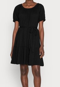Eine Person trägt ein knielanges schwarzes Kleid mit Puffärmeln und einem gebundenen Taillengürtel und steht vor einem einfarbigen Hintergrund.