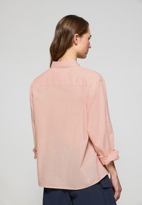 Camicia color rosa chiaro con colletto e maniche arrotolate, realizzata in un tessuto morbido e testurizzato. Presenta una spalla posteriore e una vestibilità comoda.